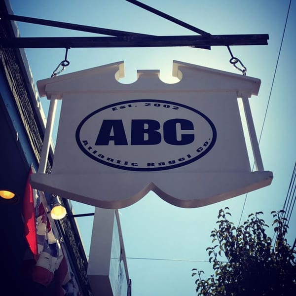 A white hanging sign for Atlantic Bagel Co. with "ABC" in a blue oval against a bright sky.