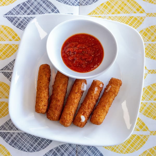 Five golden-brown mozzarella sticks with marinara sauce on a white plate, on a patterned tablecloth.