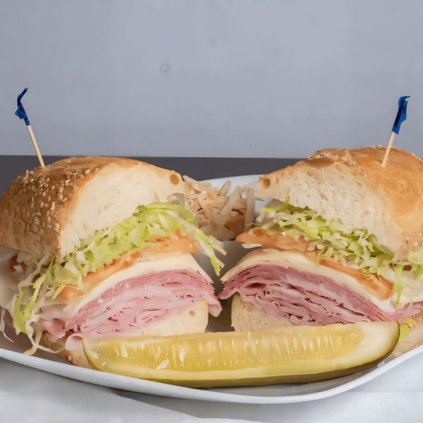 Two halves of a large deli sandwich with meat, cheese, lettuce, and tomato, with a pickle spear.