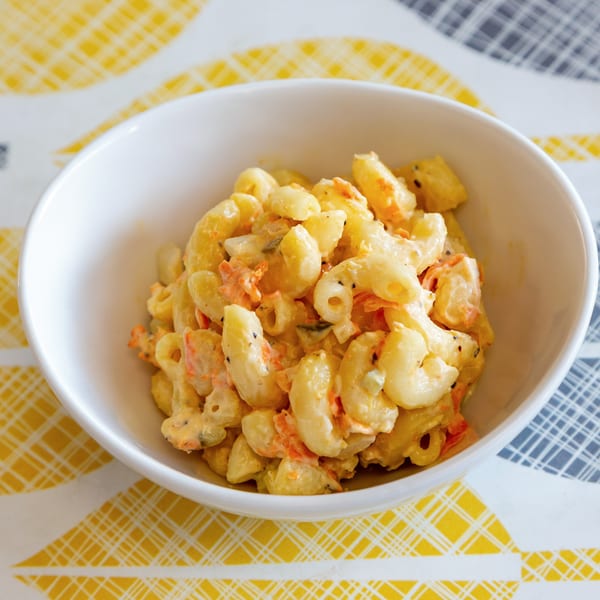 A white bowl filled with creamy macaroni salad, featuring elbow pasta and shredded carrots, on a patterned tablecloth.