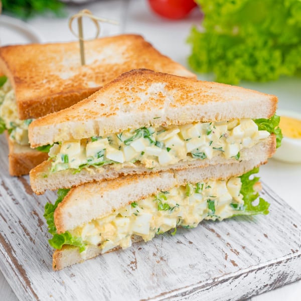 Two egg salad sandwiches on toasted bread with lettuce, cut in half, on a white cutting board.