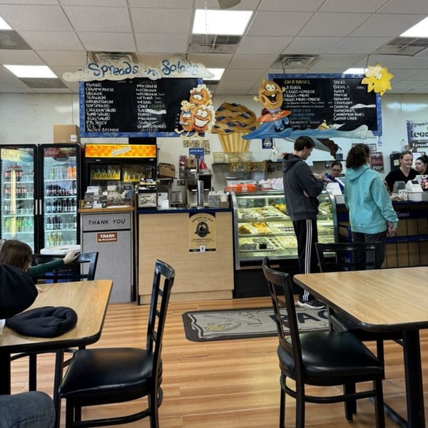 Customers order at the counter of Atlantic Bagel Co., with menu boards and display cases visible.