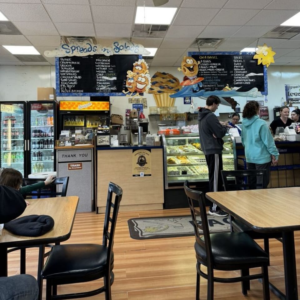 Customers order at the counter of Atlantic Bagel Co., with menu boards and display cases visible.